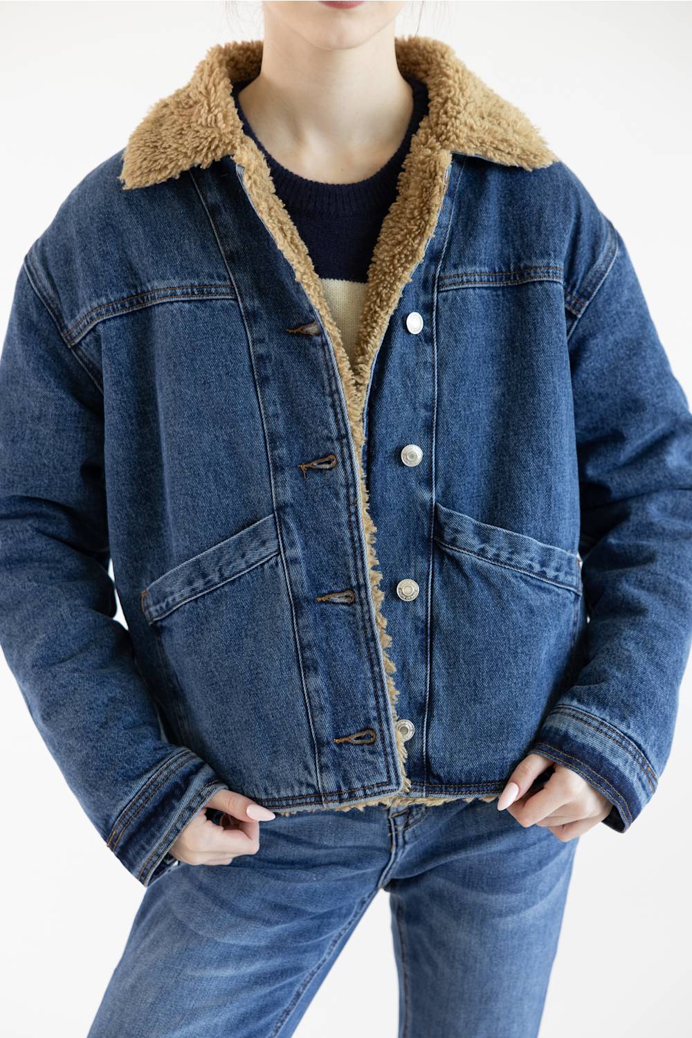 A person wearing a blue denim jacket with a tan sherpa fleece collar and lining, paired with blue jeans, against a plain white background.