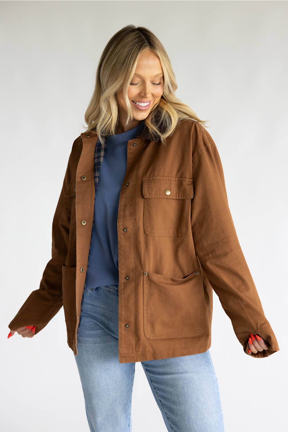 A woman wearing a brown utility jacket over a blue top and light blue jeans, standing against a light gray studio background.
