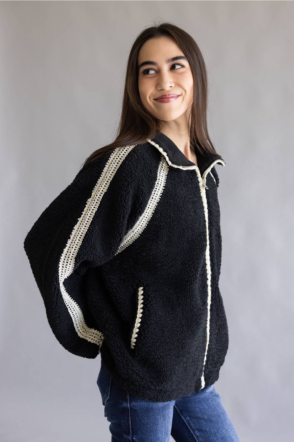 A woman wearing a black sherpa fleece zip-up jacket with cream-colored crochet trim details, paired with blue jeans, photographed against a neutral gray studio background.