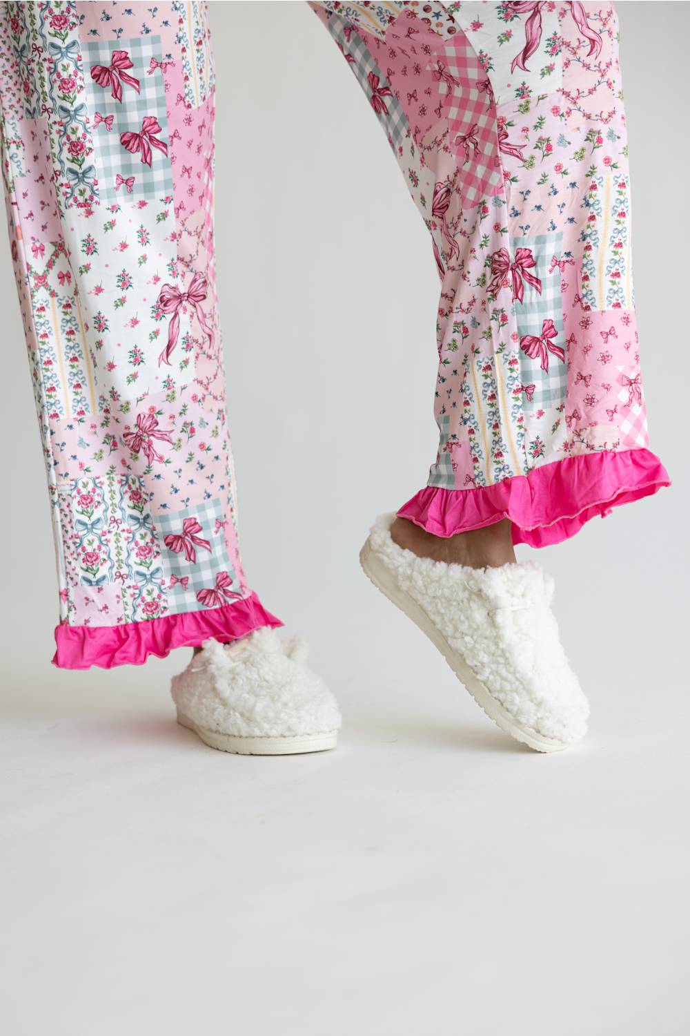 A person wearing pink patchwork pajama pants with floral and bow patterns, bright pink ruffled hems, and fluffy white slip-on slippers against a plain light background.