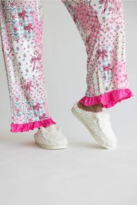 A person wearing pink patchwork pajama pants with floral and bow patterns, bright pink ruffled hems, and fluffy white slip-on slippers against a plain light background.