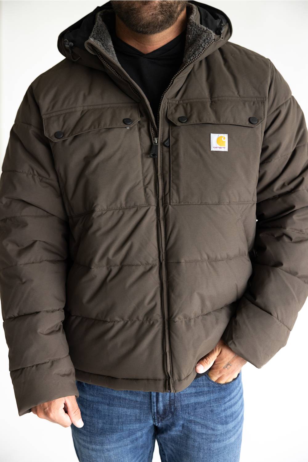 A man wearing a dark brown Carhartt puffer jacket with a hood, black shirt, and blue jeans, shown from the chin down against a white background.