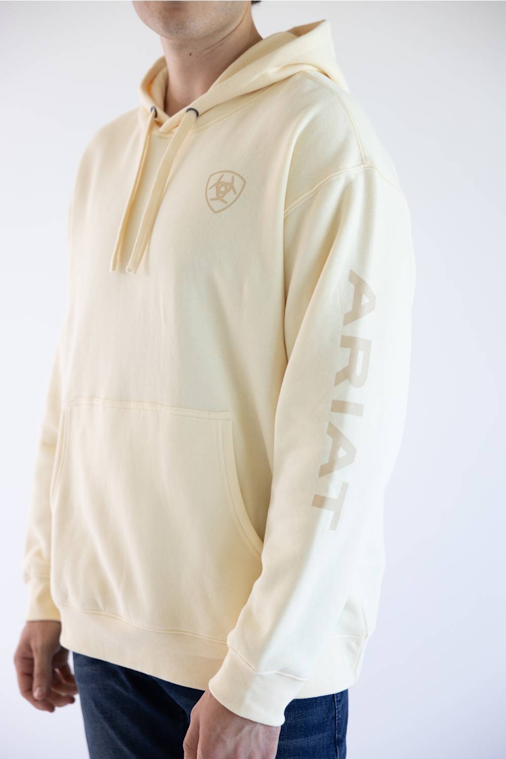 A person wearing a cream-colored hoodie with a tonal embroidered shield logo on the left chest and the word 'ARIAT' printed vertically on the right sleeve, paired with dark blue jeans against a plain white background.