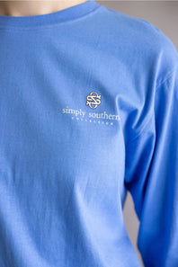 A close-up of a person wearing a long-sleeve crewneck t-shirt in a medium blue color. The shirt features a small white embroidered logo and the text 'simply southern COLLECTION' on the left chest.