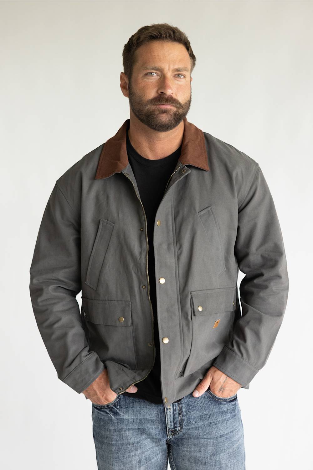 A man wearing a gray waxed cotton utility jacket with a brown corduroy collar, styled over a black t-shirt and blue jeans, standing against a light gray studio background.