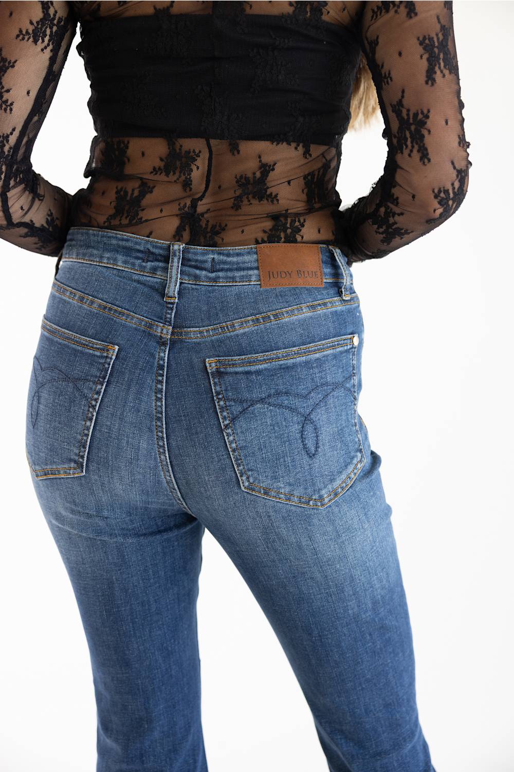 Rear view of a person wearing blue denim jeans with a brown leather patch on the back waistband and a sheer black floral long-sleeve top, against a plain white background.