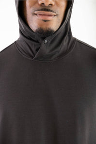 Close-up shot of a model wearing a dark raven black hooded sweatshirt featuring a structured crossover neckline with a snap button closure, cross-stitch accent below the collar, and a raised hood.