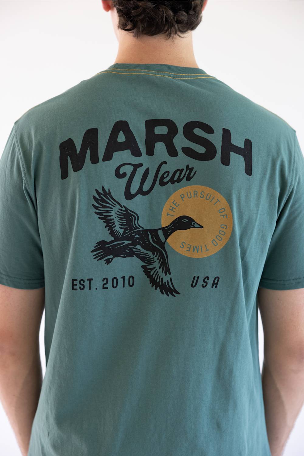 A man viewed from behind wearing a teal green t-shirt with a large black graphic on the back reading 'MARSH Wear' and 'EST. 2010 USA', featuring a flying duck and a circular gold emblem.