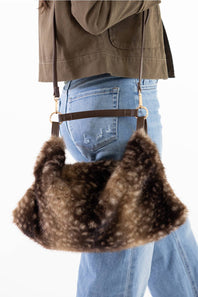 A brown faux fur shoulder bag with a dark brown leather strap and gold-tone hardware, shown being worn over the shoulder of a person wearing blue jeans and an olive green jacket.