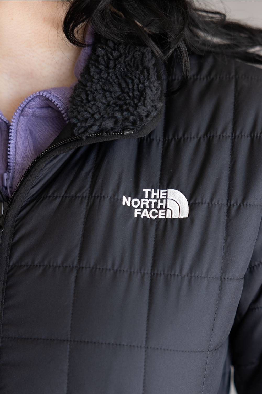 Close-up of a black puffer jacket with a white The North Face logo on the chest, featuring a dark gray sherpa collar and a purple inner lining.