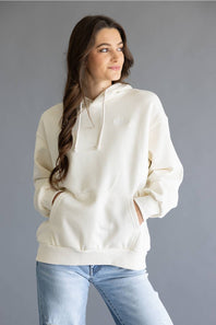 A woman wearing an oversized cream-colored hoodie with a small tonal embroidered logo on the chest, paired with light blue jeans, set against a solid gray studio background.