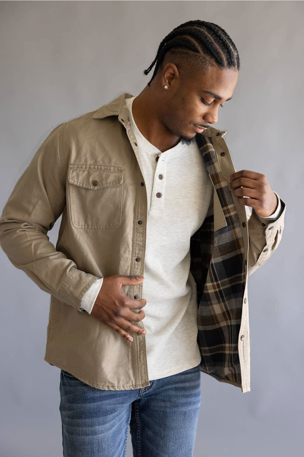 A man wearing a beige button-up overshirt with a plaid lining over a white henley shirt and blue jeans, standing against a neutral gray background.