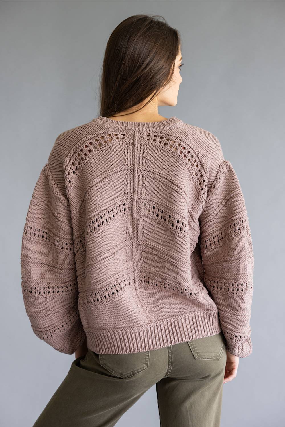 A woman shown from the back wearing a dusty pink open-knit sweater with textured chevron patterns and olive green pants, set against a solid gray studio background.