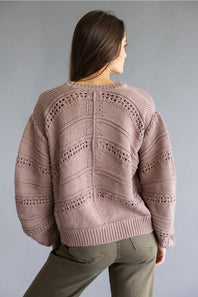 A woman shown from the back wearing a dusty pink open-knit sweater with textured chevron patterns and olive green pants, set against a solid gray studio background.