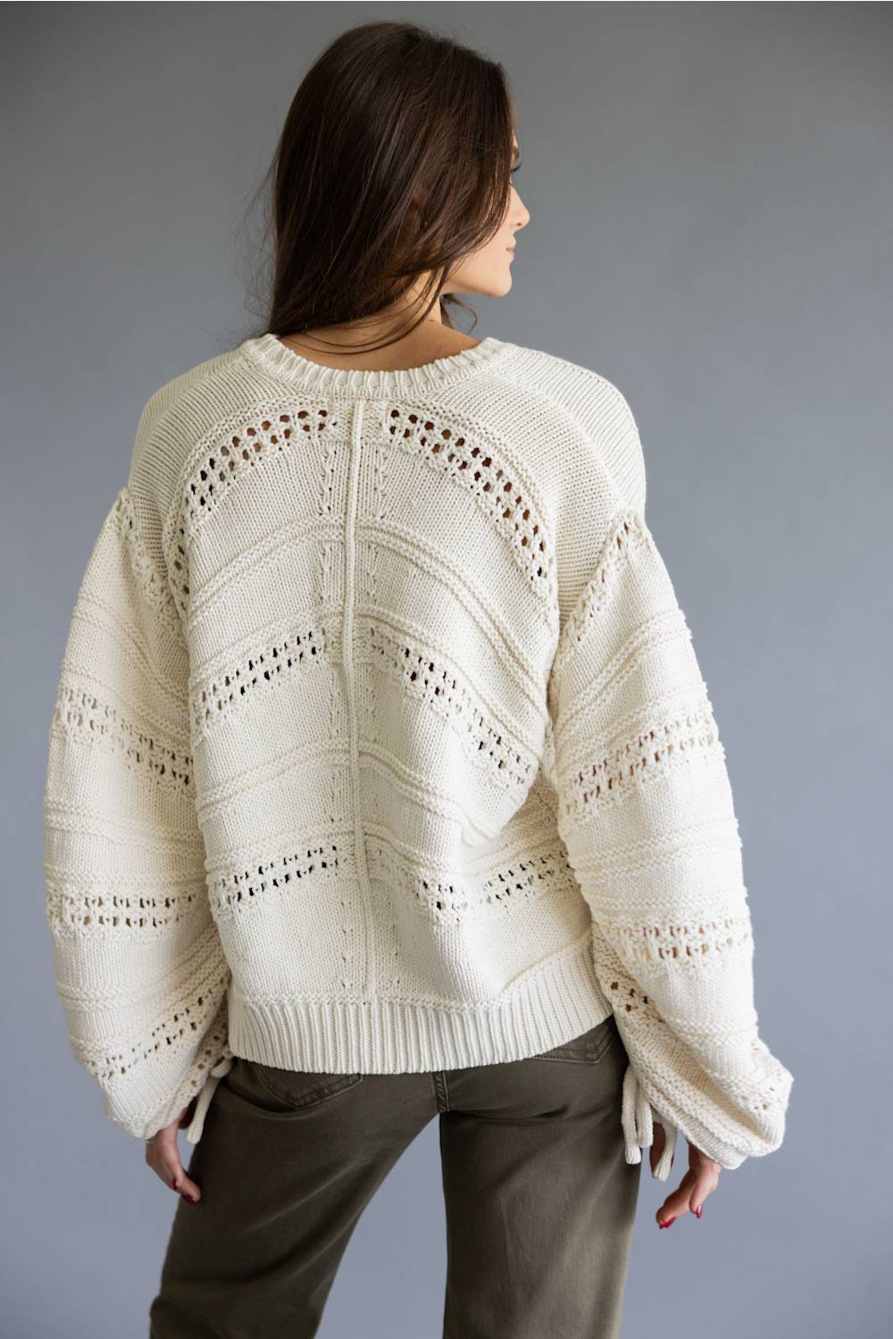 A woman shown from the back wearing a cream-colored, chunky knit sweater with intricate openwork patterns and voluminous sleeves, paired with olive green pants, against a solid gray studio background.