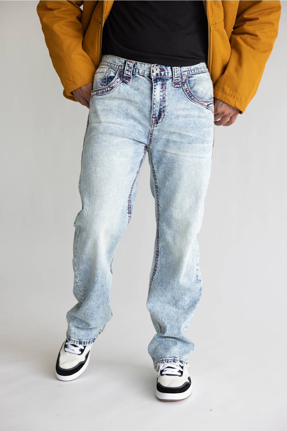 A person wearing light-wash, baggy blue jeans with a faded wash, styled with a black t-shirt, a mustard yellow jacket, and black-and-white sneakers, photographed against a plain light gray studio background.