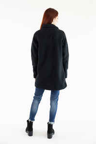 A woman viewed from behind wearing a dark charcoal black fuzzy mid-length coat, blue jeans, and black ankle boots against a plain white studio background.
