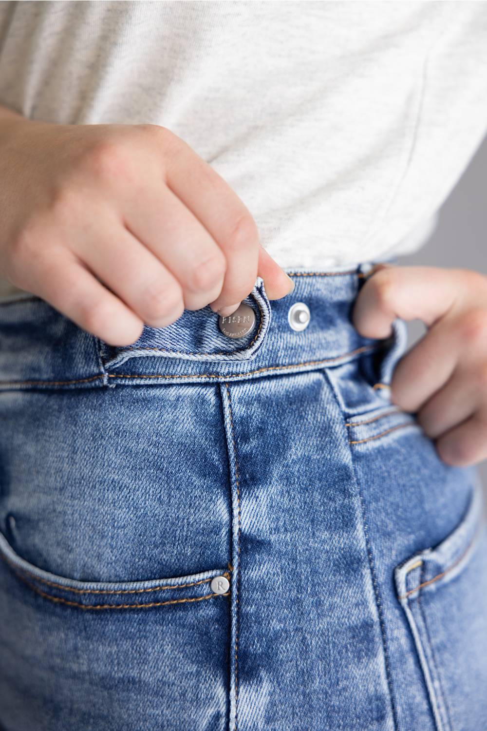 Close-up of a person's hands adjusting the waistband and button of blue denim jeans, wearing a light gray heathered t-shirt.