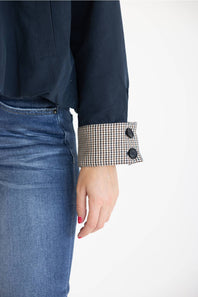 Close-up of a person wearing a dark navy long-sleeve top with a black-and-white gingham buttoned cuff, paired with blue jeans against a plain white background.