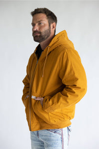 A man wearing a mustard yellow hooded jacket with a front zipper and drawstrings, paired with light blue jeans, photographed against a plain light gray studio background.