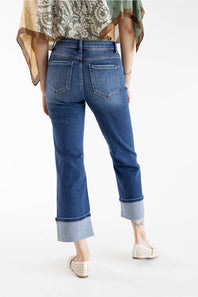 A person shown from the back wearing blue cropped jeans with rolled cuffs, a patterned blouse, and woven flat shoes against a clean white studio background.