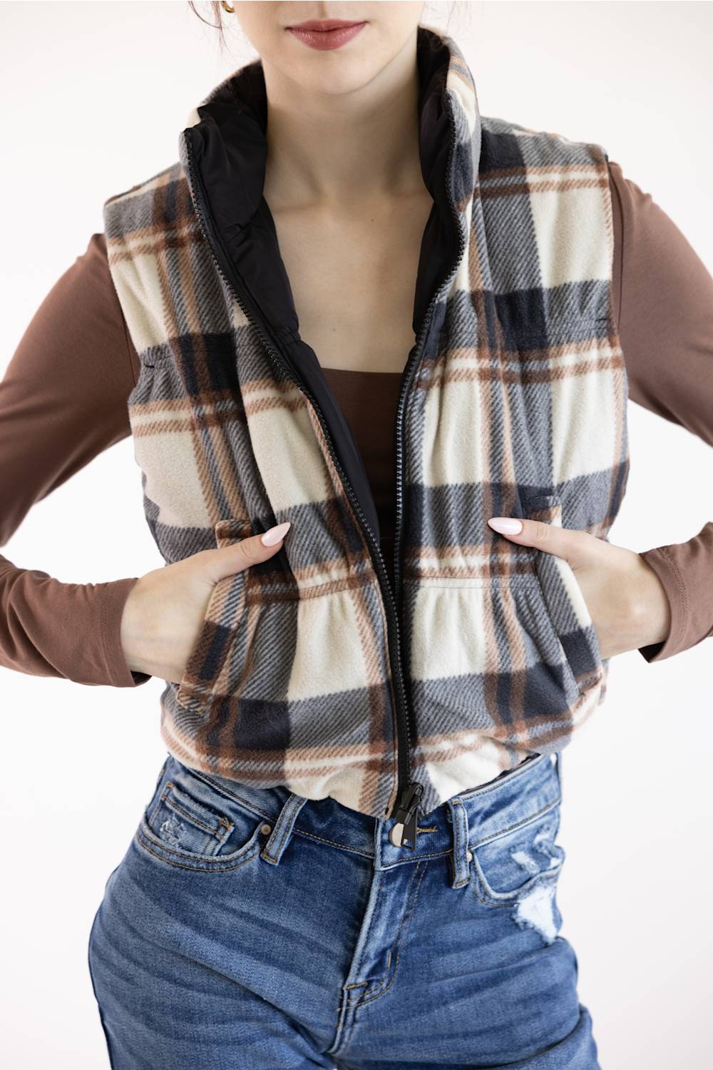 A woman wearing a zip-front plaid vest over a brown long-sleeve top and blue jeans, shown from the lower face to the hips against a plain light background.