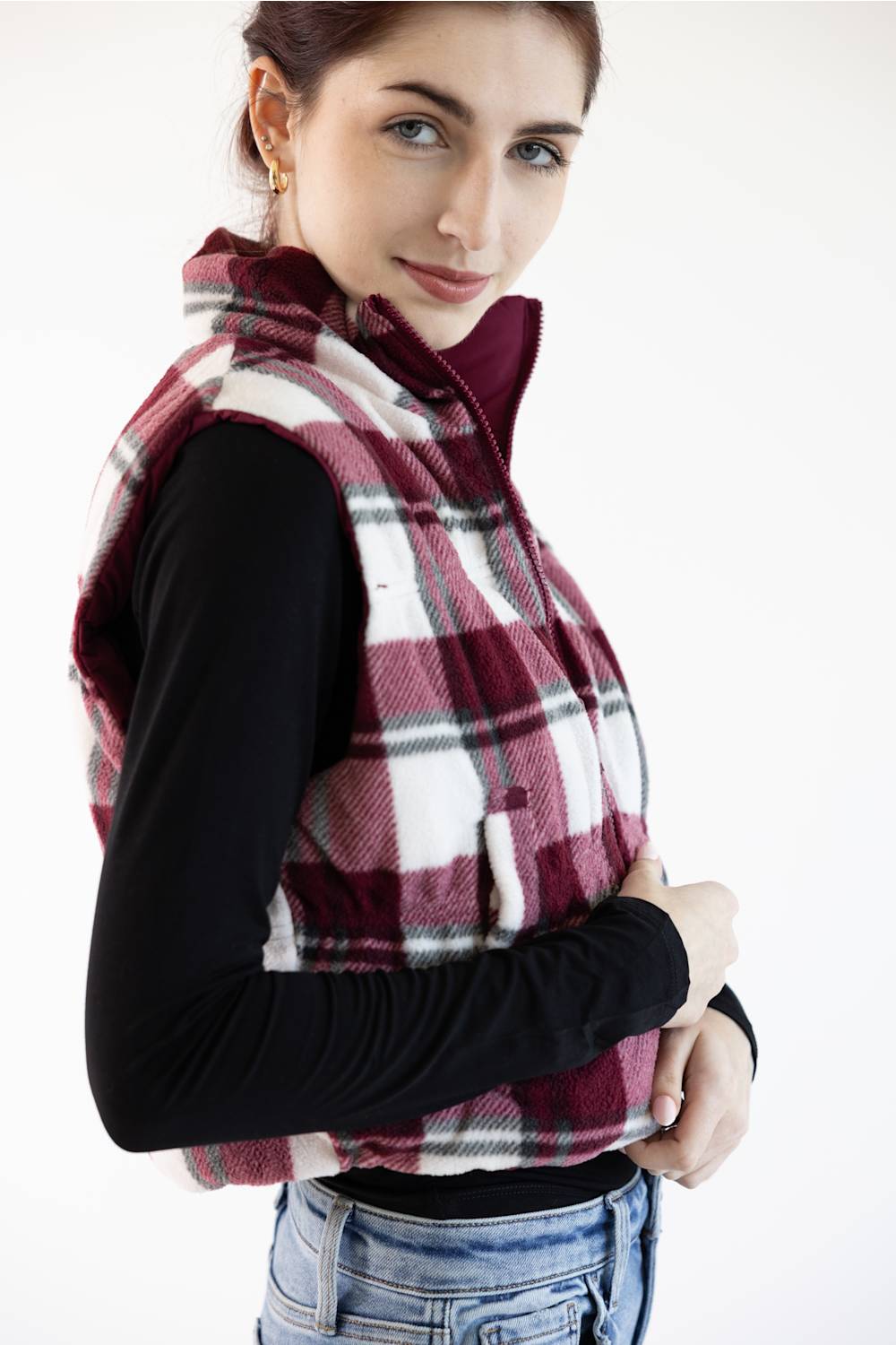 A woman wearing a burgundy, white, and gray plaid zip-up fleece vest over a black long-sleeve shirt and blue jeans, photographed against a plain white background.