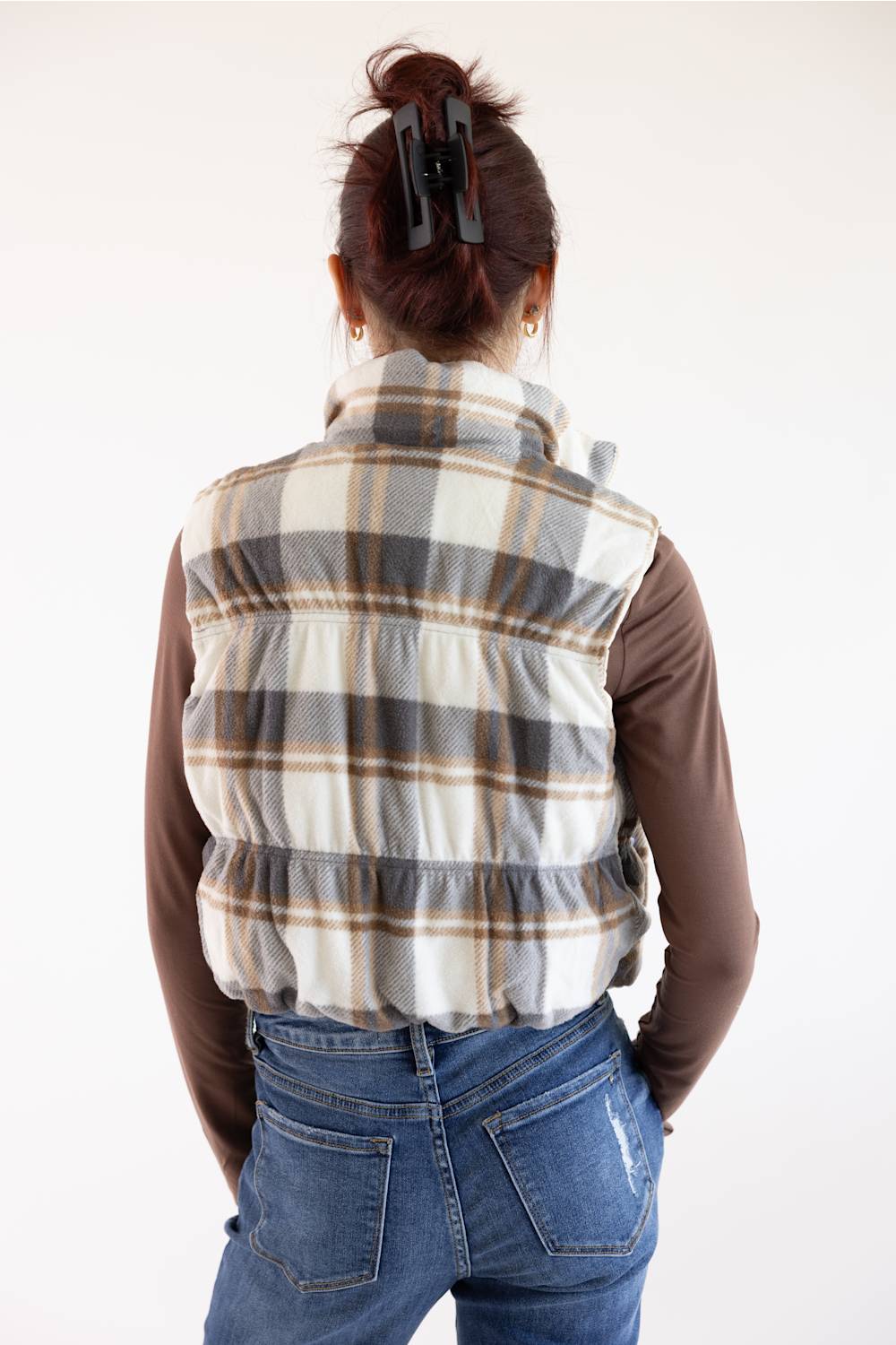 A woman viewed from behind wearing a cream, gray, and brown plaid puffer vest over a brown long-sleeve shirt and blue jeans, against a plain white background.