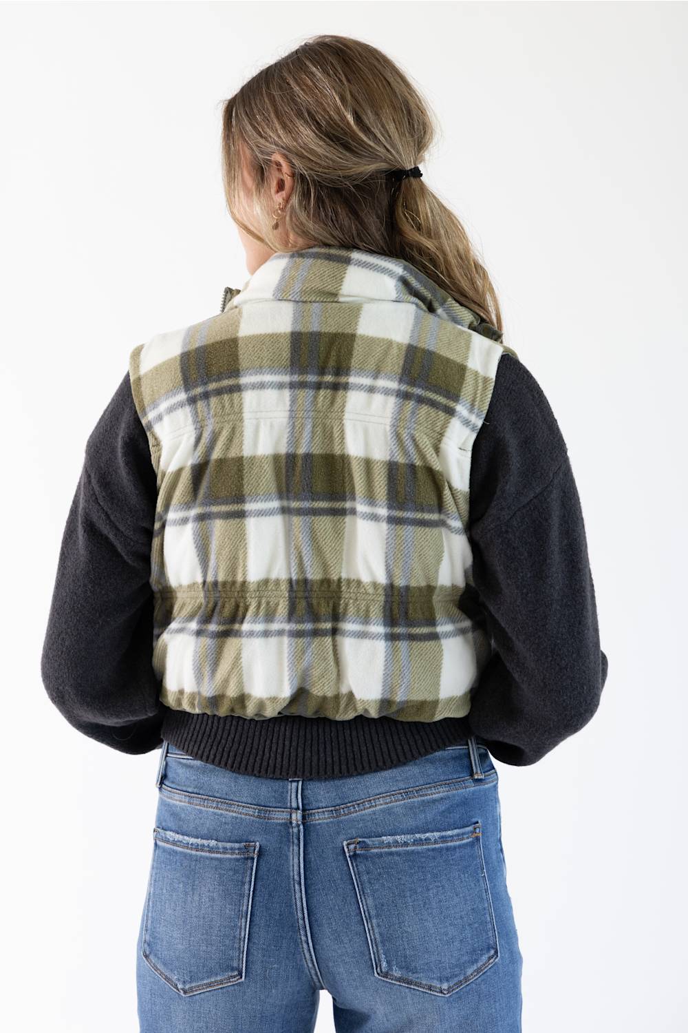 A person shown from the back wearing a sleeveless plaid quilted vest over a dark long-sleeve top and blue jeans, against a plain white studio background.