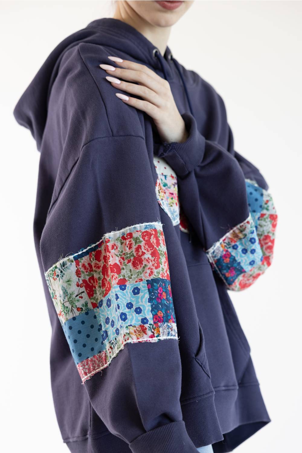 A person wearing an oversized navy hoodie with colorful patchwork floral and polka-dot sleeve panels, photographed against a plain light background.