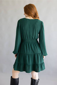 A woman modeling a dark green tiered mini dress with long balloon sleeves and black knee-high boots, standing against a neutral gray studio backdrop.