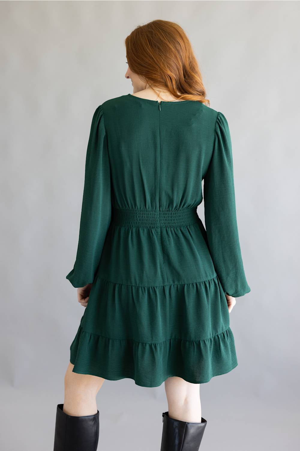 A woman modeling a dark green tiered mini dress with long balloon sleeves and black knee-high boots, standing against a neutral gray studio backdrop.