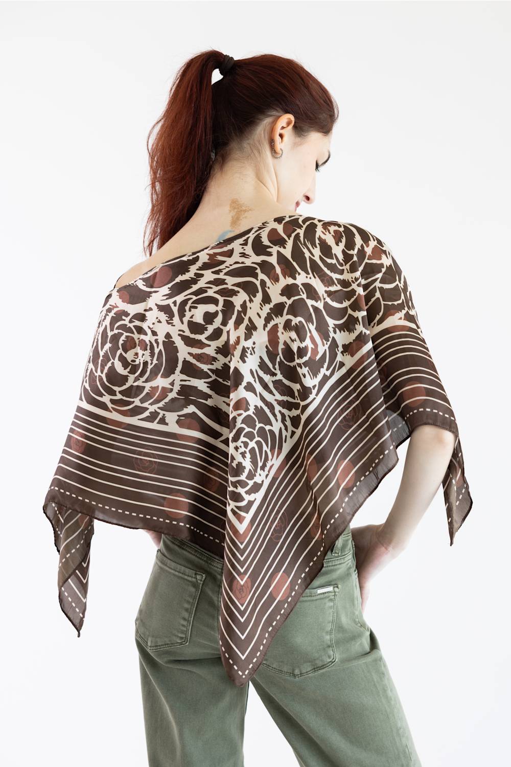 A woman with red hair tied back is shown from behind wearing a brown and beige patterned scarf draped over her shoulders, paired with olive green pants, against a plain white background.