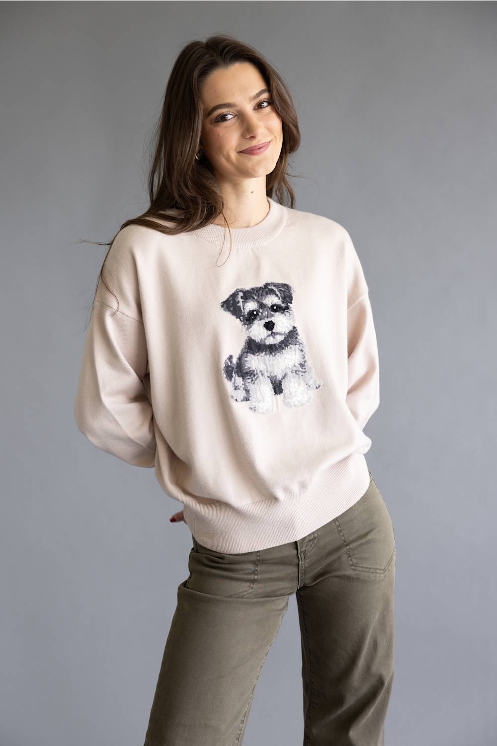A woman wearing a light beige crewneck sweatshirt with a black and white dog graphic on the front, paired with olive green pants, against a solid gray studio background.