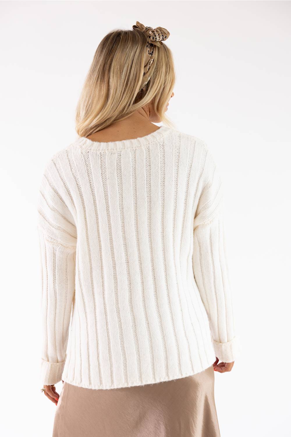 A woman viewed from the back wearing a cream ribbed knit sweater and a beige skirt against a plain white studio background.