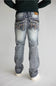 Rear view of a person wearing light-wash blue denim jeans with heavily embellished decorative back pockets, a dark green long-sleeve top, and white sneakers, set against a plain light gray studio background.