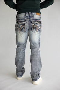 Rear view of a person wearing light-wash blue denim jeans with heavily embellished decorative back pockets, a dark green long-sleeve top, and white sneakers, set against a plain light gray studio background.