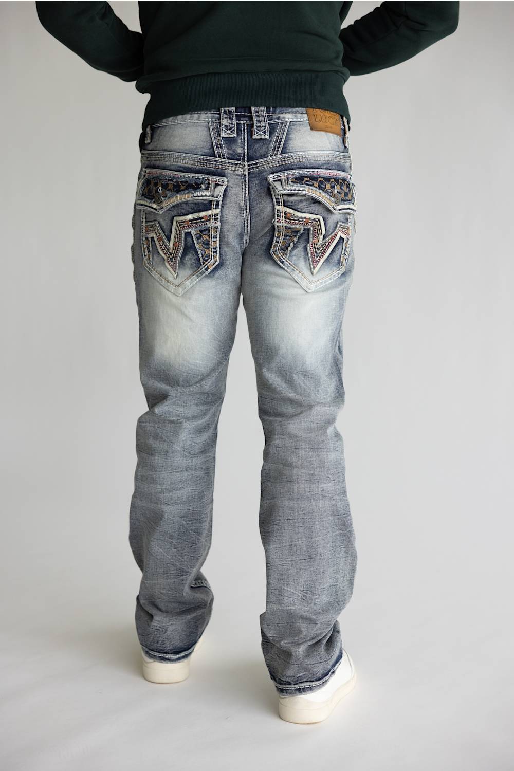 Rear view of a person wearing light-wash blue denim jeans with heavily embellished decorative back pockets, a dark green long-sleeve top, and white sneakers, set against a plain light gray studio background.