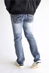 Rear view of a person wearing light-wash blue jeans with decorative embroidered detailing on the back pockets, paired with a black jacket and white sneakers, against a plain white studio background.