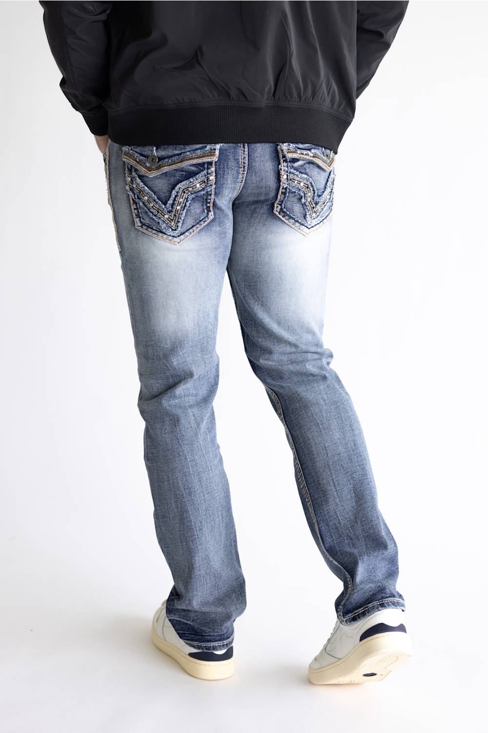 Rear view of a person wearing light-wash blue jeans with decorative embroidered detailing on the back pockets, paired with a black jacket and white sneakers, against a plain white studio background.