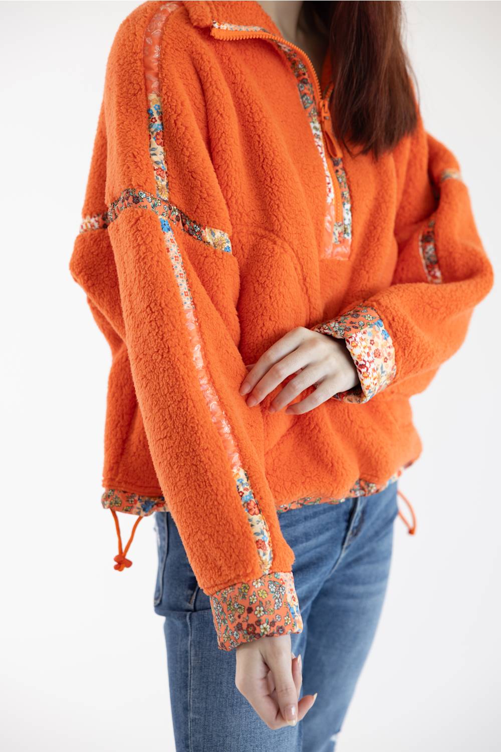 An orange sherpa fleece pullover sweater with floral patterned trim on the collar, cuffs, and front seams, worn by a model with blue jeans against a white background.
