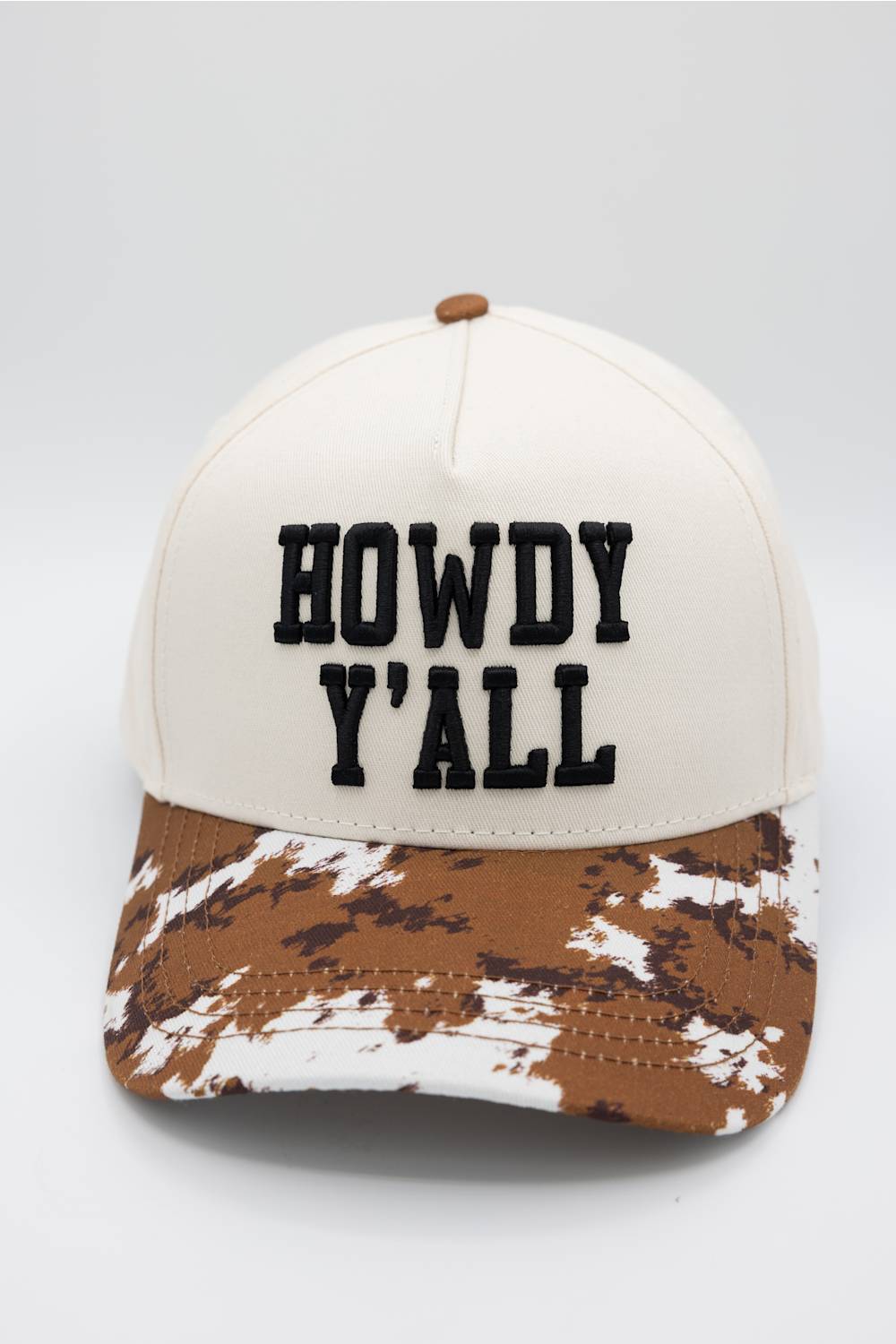 A baseball cap with an off-white crown and a brown and white camouflage patterned brim. The front features the text 'HOWDY Y'ALL' embroidered in black block letters.
