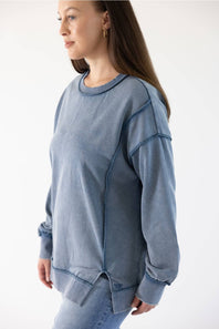A woman wearing a washed slate blue crewneck sweatshirt with unique diagonal seam details, paired with light blue jeans, shown from a side profile against a plain white background.