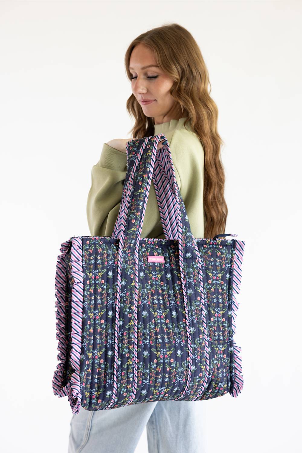 A woman wearing a sage green top and light blue jeans holds a large tote bag. The bag has a navy blue base with a colorful floral pattern and purple fringe details along the edges and handles.