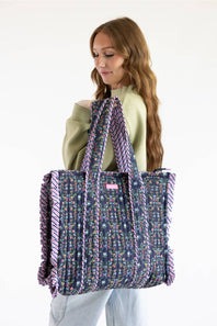 A woman wearing a sage green top and light blue jeans holds a large tote bag. The bag has a navy blue base with a colorful floral pattern and purple fringe details along the edges and handles.