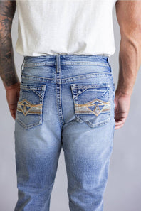 Rear view of a person wearing light blue denim jeans with decorative embroidered patch detailing on the back pockets, paired with a white t-shirt against a neutral gray background.