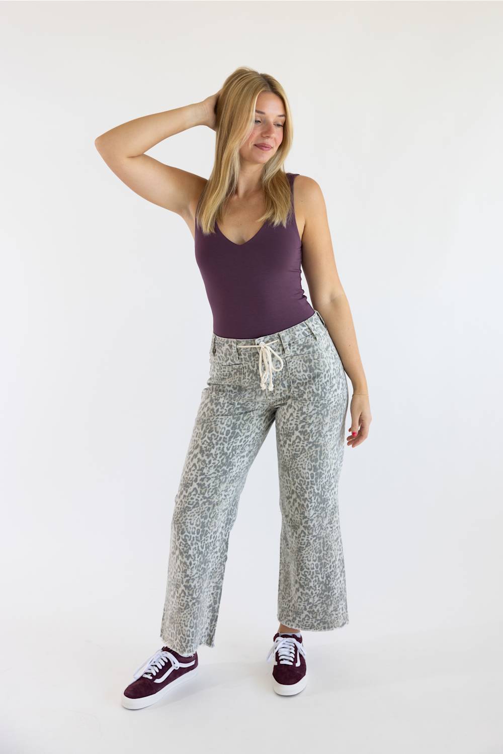 A woman modeling a pair of wide-leg, cropped gray and white patterned sweatpants with a white drawstring, paired with a purple tank top and burgundy sneakers, against a plain white studio background.