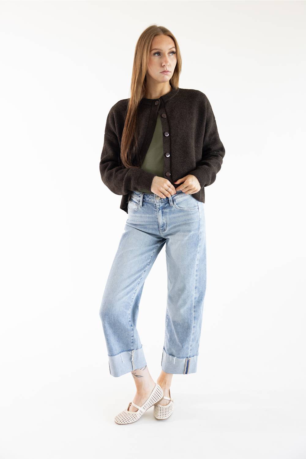 A woman modeling a dark brown knit button-up cardigan over an olive green top, paired with light-wash blue cuffed jeans and beige flat shoes, against a plain white studio background.