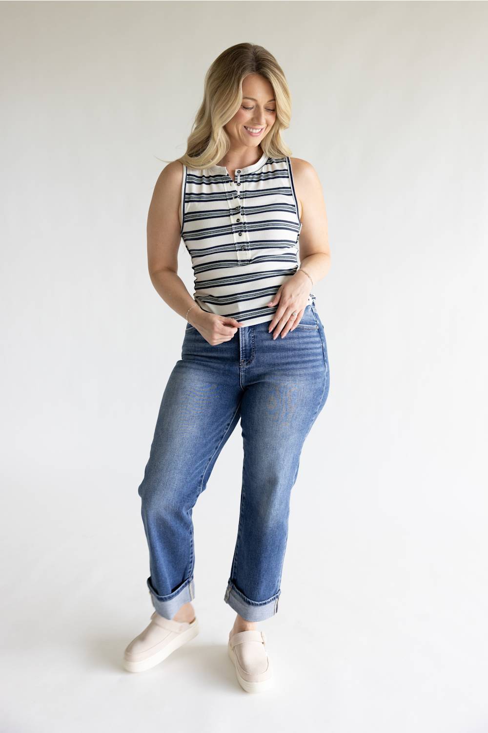 A woman modeling a sleeveless top with navy and white horizontal stripes, paired with blue jeans and off-white slip-on shoes, against a plain light gray studio background.