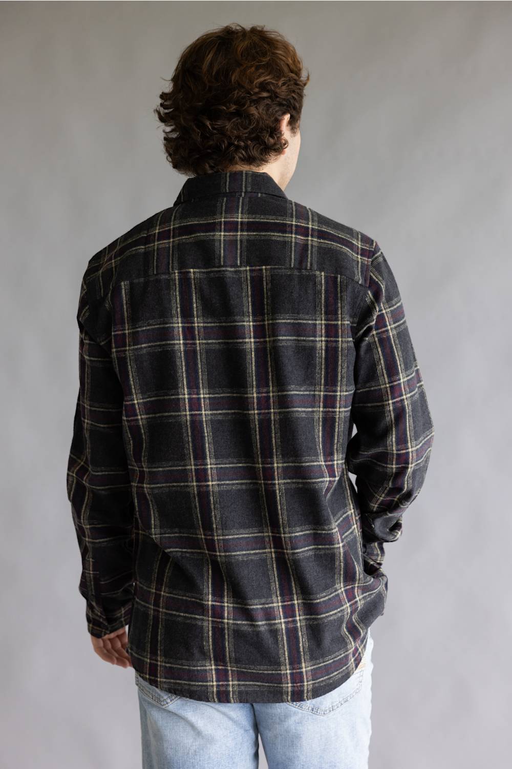 Back view of a male model wearing the Split Flannel Shirt for Men in Slate, showcasing the charcoal gray plaid pattern with burgundy and cream accent lines, straight back yoke, long sleeves, and rounded hem against a plain gray studio background.